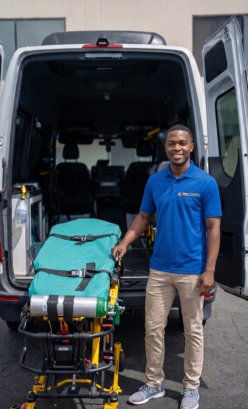 A smiling man in a blue polo shirt stands beside a stretcher at the open back of an ambulance van, conveying readiness and professionalism.